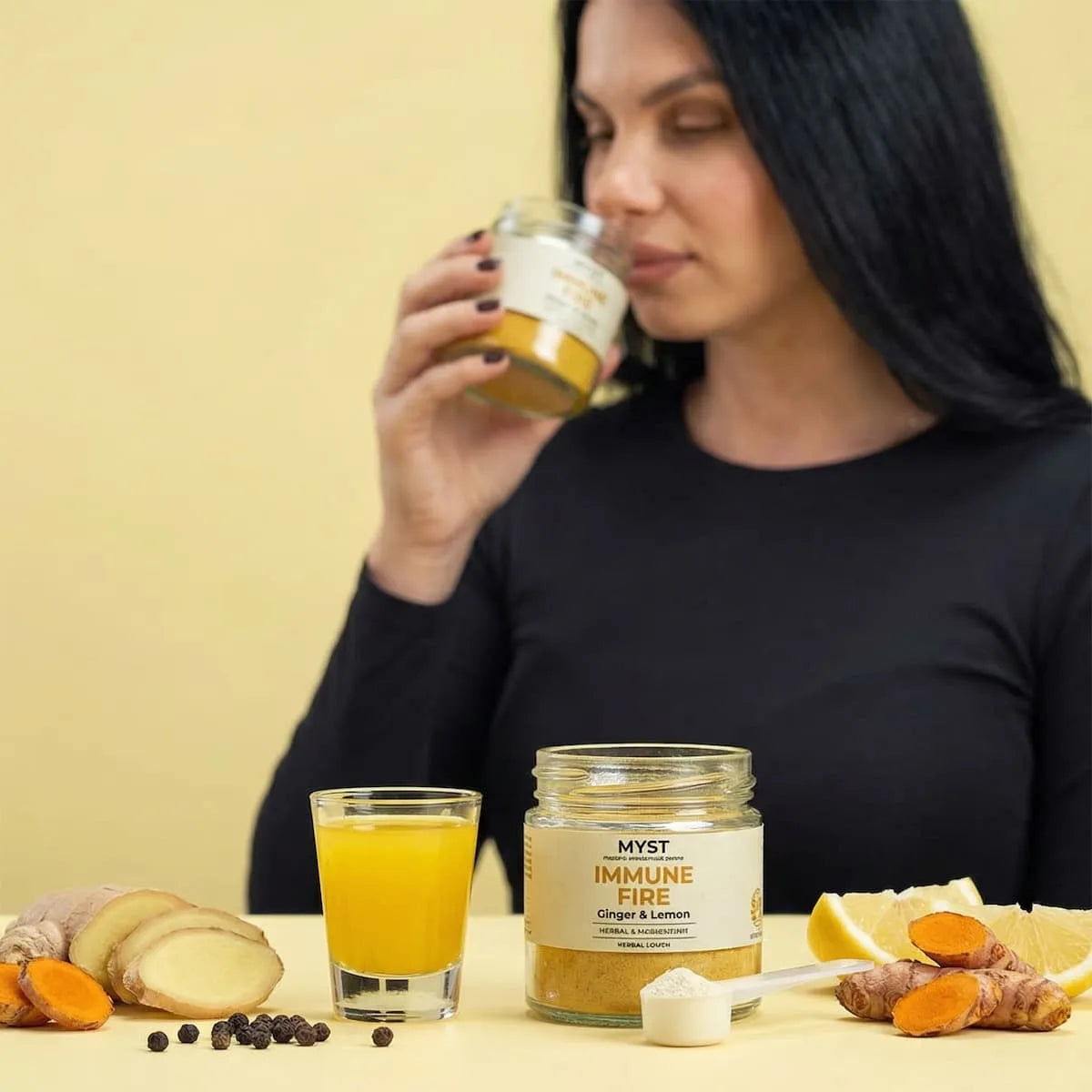 Woman holding a jar of 'MYST Immune Fire' with a glass of orange juice and ingredients on a table.