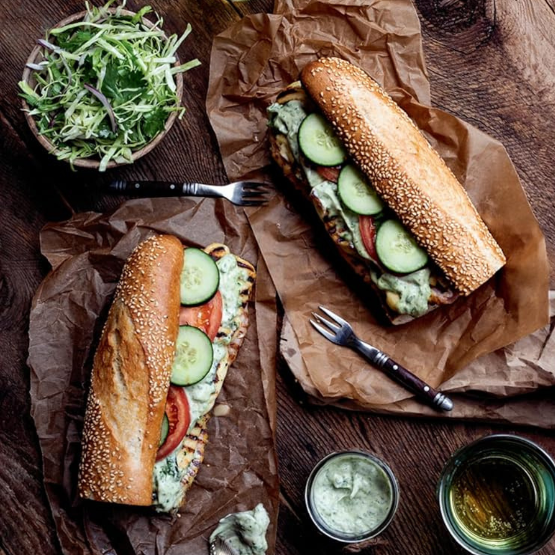 Two sub sandwiches with cucumbers and tomatoes on a wooden table with condiments.