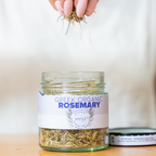 Jar of Greek organic rosemary with a hand holding rosemary sprigs above it on a light background.