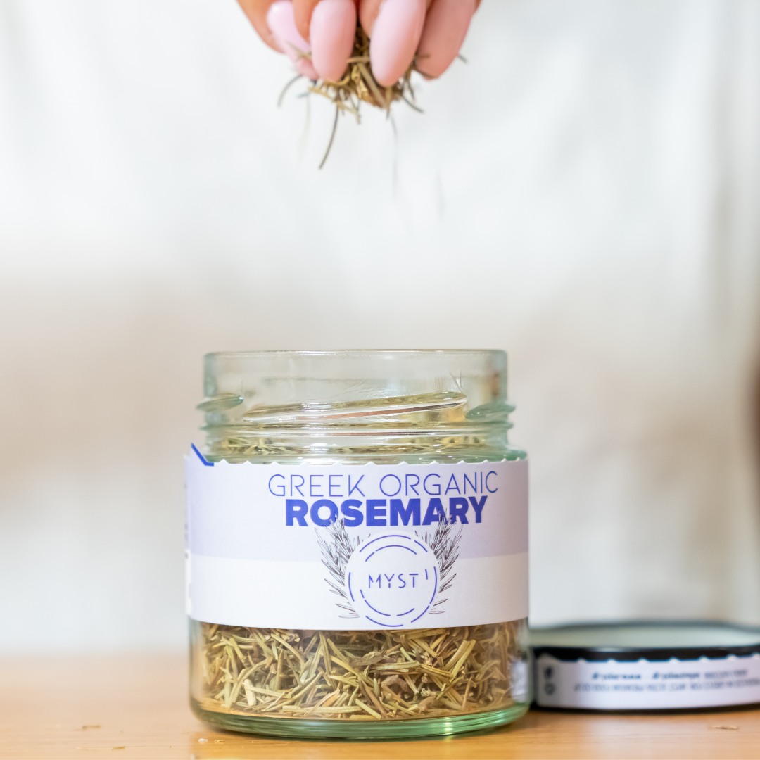 Jar of Greek organic rosemary with a hand holding rosemary sprigs above it on a light background.