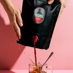 Person pouring coffee from a black bag labeled 'Cold Brew Collective' into a glass mug on a pink background.