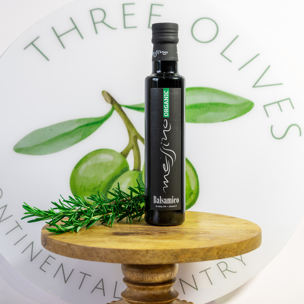 Bottle of Three Olives Organic Balsamicio on a wooden stand with green olives and rosemary.