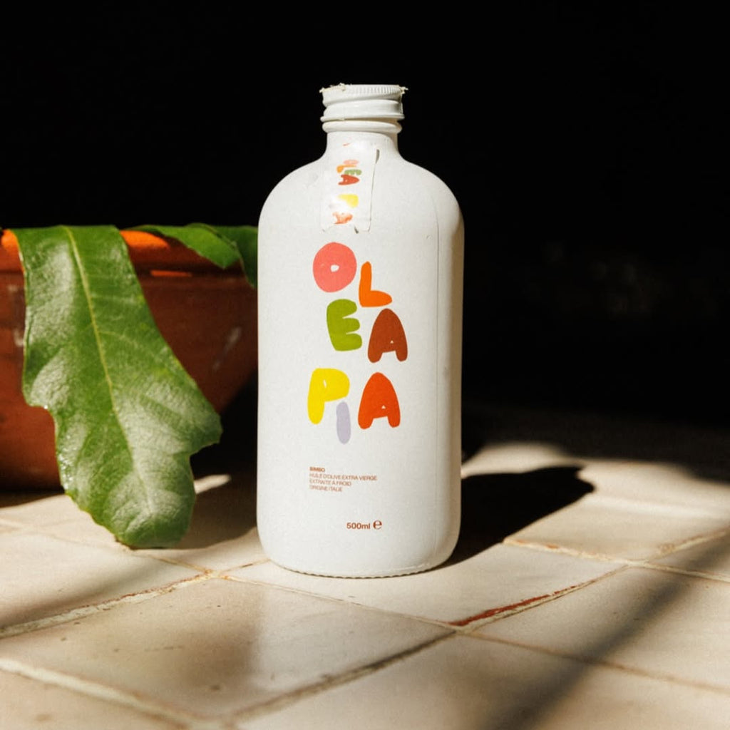 White bottle with colorful 'Olea' branding on a tiled surface with a leaf in the background