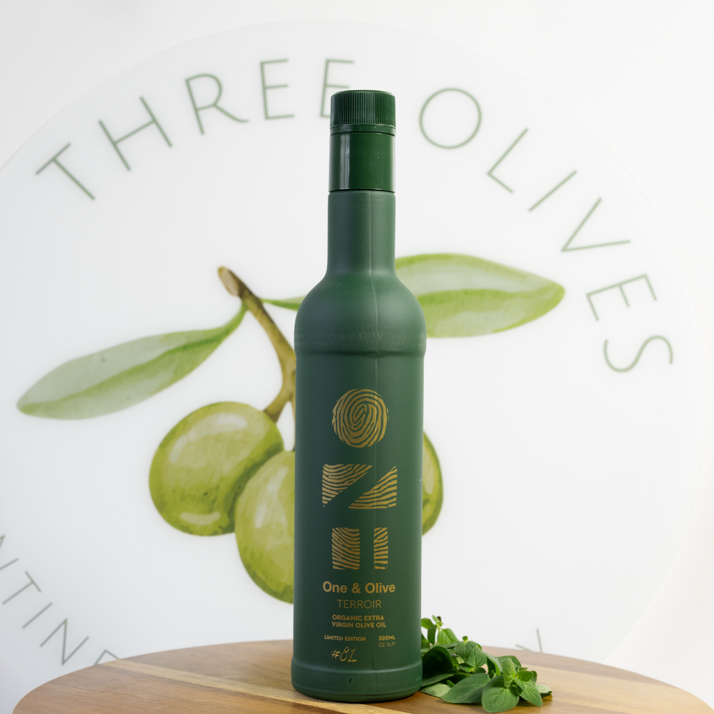 Green bottle labeled 'One & Olive' on a wooden surface with olives and leaves in the background.