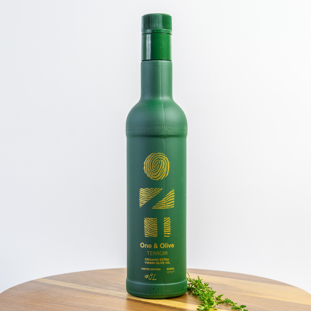 Green bottle of One & Olive olive oil on a wooden surface with a white background