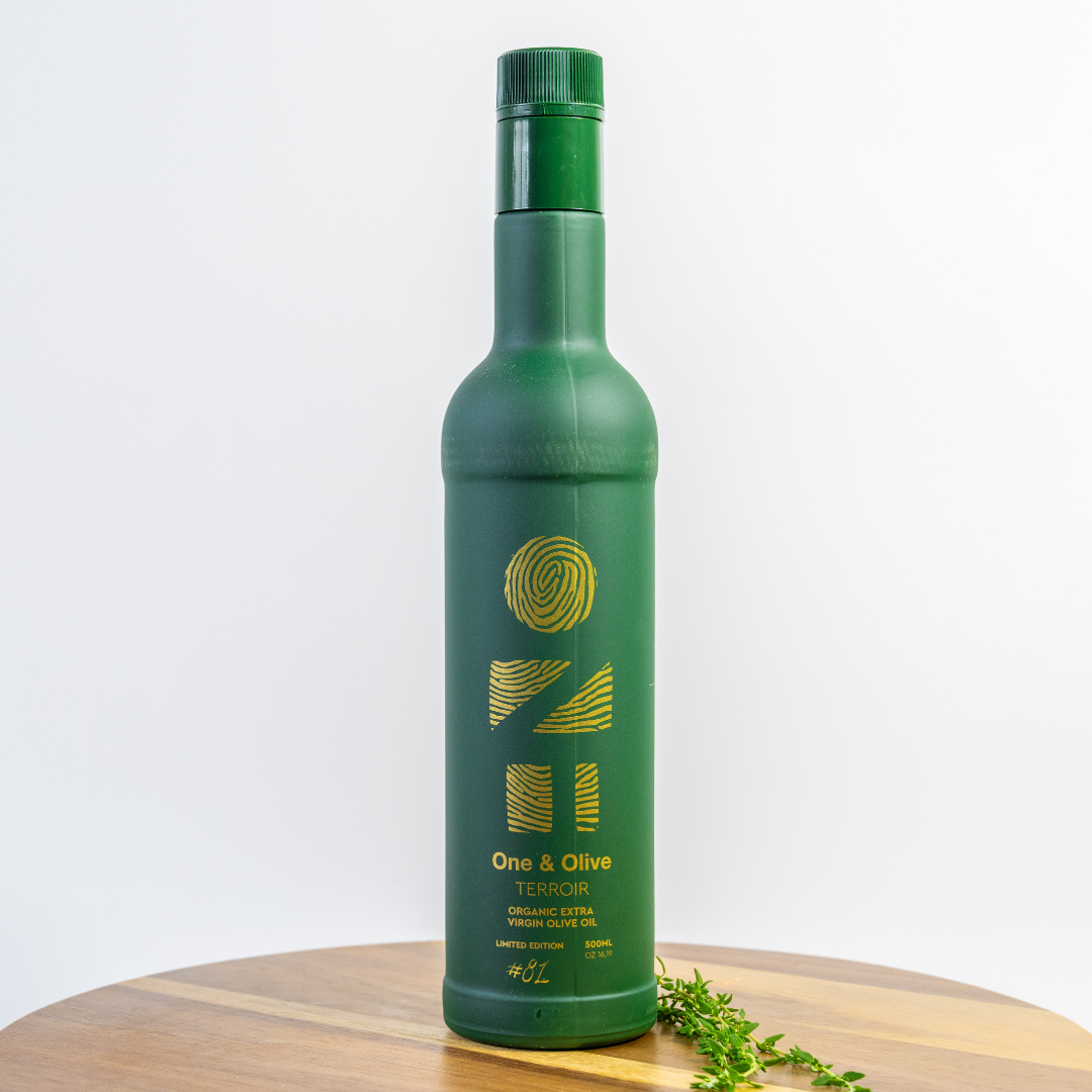 Green bottle of One & Olive olive oil on a wooden surface with a white background