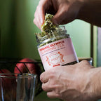 Person adding dried herbs from a jar labeled 'Organic Olympus Slimming Tea' into a glass.