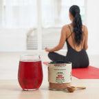Red detox supplement with a glass of red liquid on a table, with a person sitting on a yoga mat in the background.